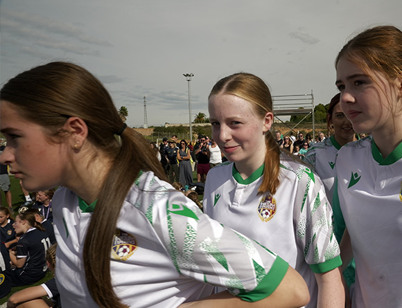three young female soccer players wearing white jerseys with green accents walking in line towards a sports event showcasing teamwork and dedication to the sport in a dynamic atmosphere celebrating youth soccer and under 12 competition