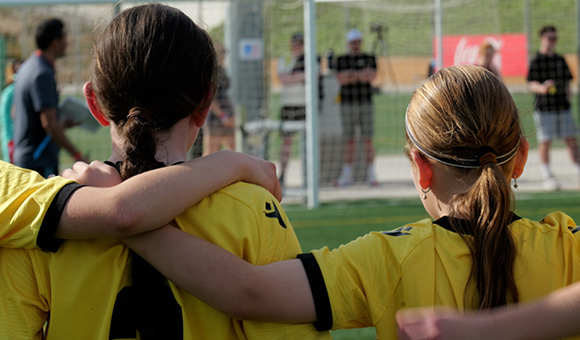 two girls in yellow soccer jerseys with arms around each other watching a game on a sunny day showing teamwork and sportsmanship in youth soccer 8 players on the field